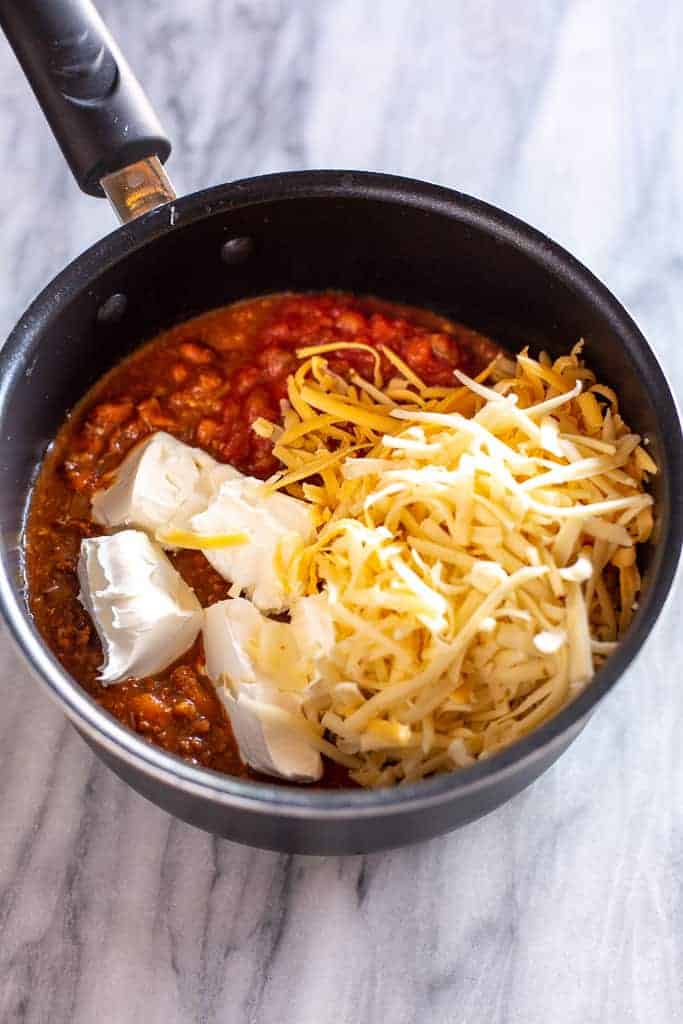 A saucepan with chili, salsa , shredded cheese and cut up cream cheese.