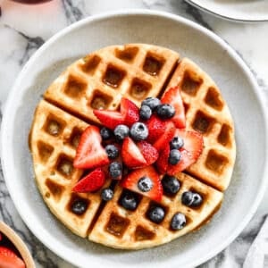 A Belgian waffle on a plate with fresh berries and syrup on top.